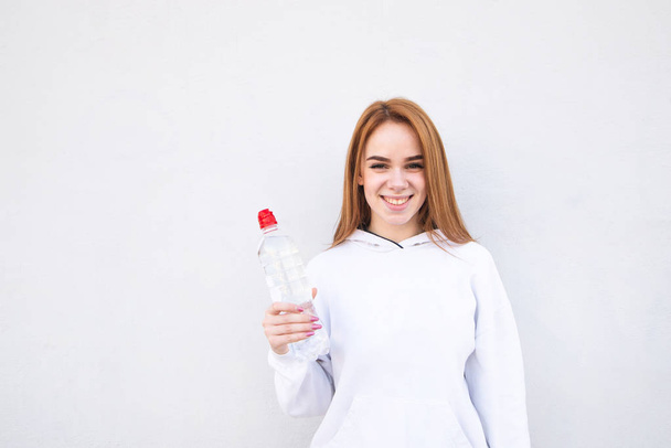 Gelukkig meisje in witte kleding met een flesje water in haar handen op een witte achtergrond glimlacht en kijkt naar de camera. Jonge aantrekkelijke atleet tegen een witte achtergrond en copyspace - Foto, afbeelding