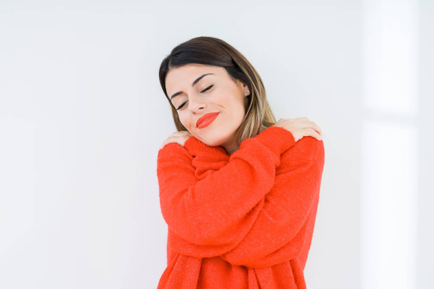 Mujer joven vistiendo suéter rojo casual sobre un fondo aislado abrazándose feliz y positivo, sonriendo confiado. Amor propio y cuidado personal
 - Foto, Imagen