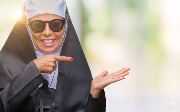 A középkorú vezető katolikus apáca nő több mint meghökkent, és mosolyog a kamerába, miközben bemutatja a kezét és ujjával mutatva elszigetelt háttér napszemüveget visel. - Fotó, kép