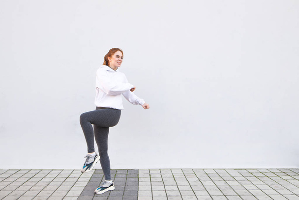 白い壁の背景でのウォーミング アップ運動少女の肖像画。朝の体操と白い背景の壁にスポーツウェアで幸せな若い女の子. - 写真・画像