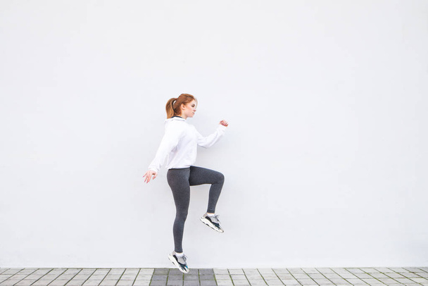 Приваблива молода дівчина стрибає на тлі білої стіни, розігрівається перед бігом і благає про спорт. Спортивна концепція. Копіспазмі
 - Фото, зображення