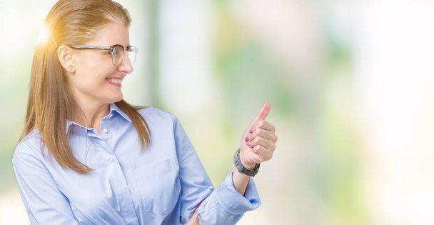 Beautiful middle age mature business woman wearing glasses over isolated background Looking proud, smiling doing thumbs up gesture to the side - Zdjęcie, obraz