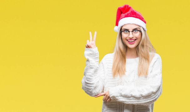 Mladá krásná blondýna žena nosí vánoční čepice izolované pozadí usmívající se smajlík, mrkl na fotoaparát dělá znamení vítězství. Číslo dvě. - Fotografie, Obrázek