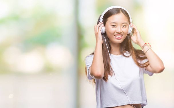 junge asiatische Frau hört Musik mit Kopfhörern isoliert Hintergrund mit einem glücklichen Gesicht stehend und lächelnd mit einem selbstbewussten Lächeln zeigt Zähne - Foto, Bild
