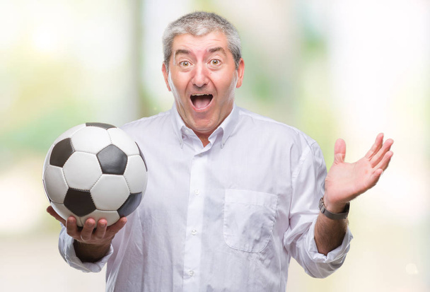 Bell'uomo anziano che tiene il pallone da calcio su sfondo isolato molto felice ed eccitato, espressione del vincitore che celebra la vittoria urlando con grande sorriso e mani alzate
 - Foto, immagini