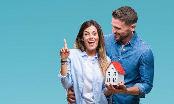 Jong koppel in liefde bedrijf huis over geïsoleerd achtergrond verrast met een idee of vraag vinger met blij gezicht, nummer één - Foto, afbeelding