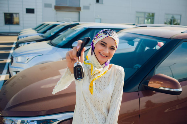 property and people concept - muslim woman in hijab with car key over car show background. happy woman taking car key from dealer in auto show or salon - Фото, зображення