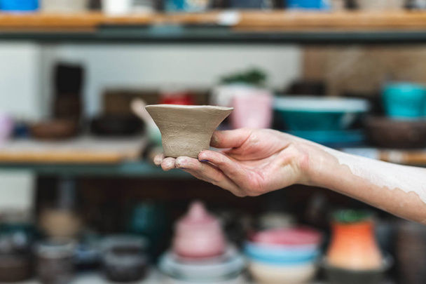 Master class concept. Cropped close up photo of workmanship lady in her workwear she stand against blurred wooden shelf background indoor workspace hold small ceramics clay tea cup on hand - Photo, Image