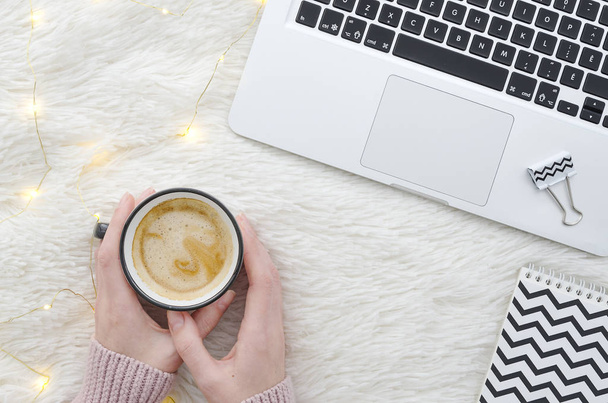 Warm cozy winter workspace background. Flat lay. Girl holding a cup of coffee on fur fluffy white background with lights garland and laptop. Top view mockup - Photo, Image