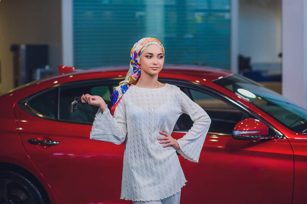 property and people concept - muslim woman in hijab with car key over car show background. happy woman taking car key from dealer in auto show or salon - Photo, image