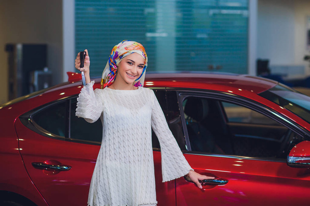 property and people concept - muslim woman in hijab with car key over car show background. happy woman taking car key from dealer in auto show or salon - 写真・画像