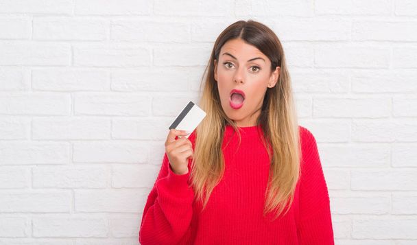 Young adult woman over white brick wall holding credit card scared in shock with a surprise face, afraid and excited with fear expression - Фото, изображение