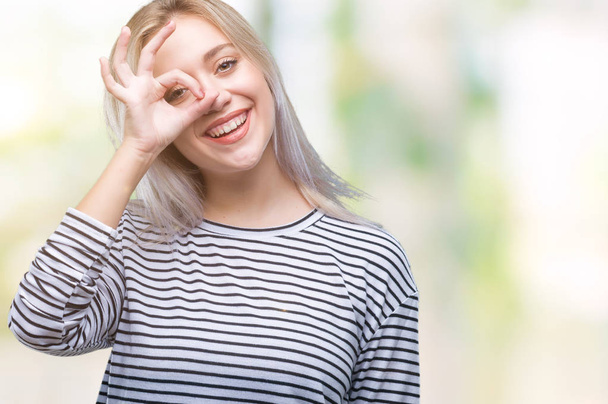 Young blonde woman over isolated background doing ok gesture with hand smiling, eye looking through fingers with happy face. - Фото, зображення