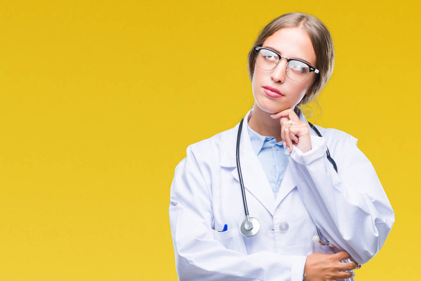 Beautiful young blonde doctor woman wearing medical uniform over isolated background with hand on chin thinking about question, pensive expression. Smiling with thoughtful face. Doubt concept. - 写真・画像