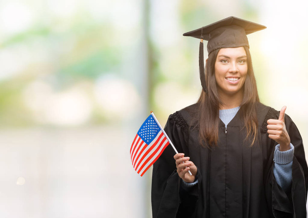 Mujer hispana joven con uniforme graduado sosteniendo la bandera de América feliz con una gran sonrisa haciendo signo de ok, pulgar hacia arriba con los dedos, excelente signo
 - Foto, Imagen