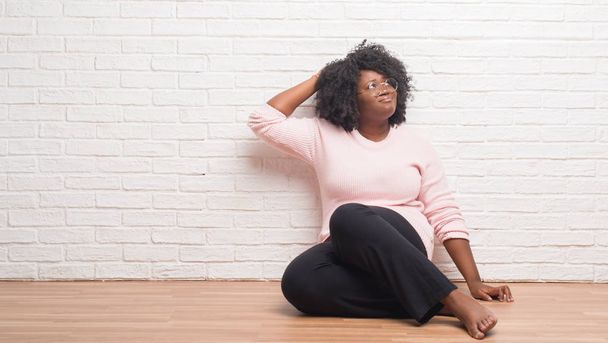 Mujer afroamericana joven sentada en el suelo en casa confunde y se pregunta acerca de la pregunta. Incierto con la duda, pensando con la mano en la cabeza. Concepto pensativo
. - Foto, imagen