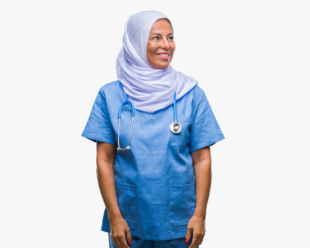 Enfermera árabe mayor de mediana edad que usa hijab sobre un fondo aislado mirando hacia otro lado con sonrisa en la cara, expresión natural. Riendo confiado
. - Foto, imagen