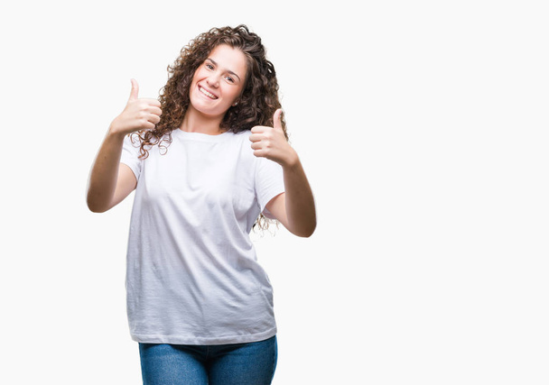 Beautiful brunette curly hair young girl wearing casual t-shirt over isolated background success sign doing positive gesture with hand, thumbs up smiling and happy. Looking at the camera with cheerful expression, winner gesture. - Fotografie, Obrázek