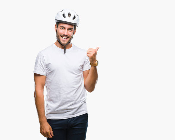 Joven hombre guapo con casco de seguridad ciclista sobre fondo aislado sonriendo con la cara feliz mirando y señalando a un lado con el pulgar hacia arriba
. - Foto, imagen