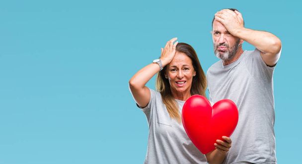 Middle age hispanic casual couple in love holding red heart over isolated background stressed with hand on head, shocked with shame and surprise face, angry and frustrated. Fear and upset for mistake. - Foto, afbeelding