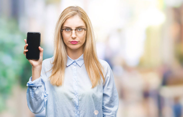 Young beautiful blonde business woman showing screen of smartphone over isolated background with a confident expression on smart face thinking serious - 写真・画像