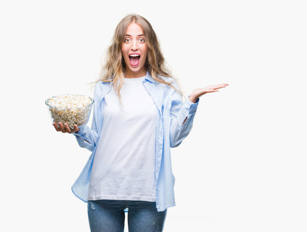 schöne junge blonde Frau isst Popcorn vor isoliertem Hintergrund sehr glücklich und aufgeregt, Siegerausdruck feiert Sieg schreiend mit breitem Lächeln und erhobenen Händen - Foto, Bild