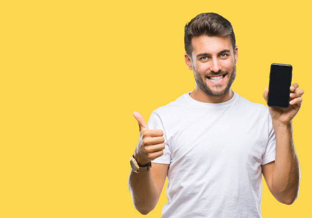 Young handsome man showing smartphone screen over isolated background happy with big smile doing ok sign, thumb up with fingers, excellent sign - Photo, Image