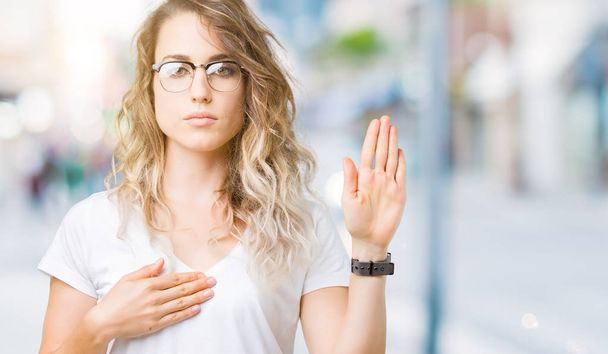Beautiful young blonde woman wearing glasses over isolated background Swearing with hand on chest and open palm, making a loyalty promise oath - Photo, Image