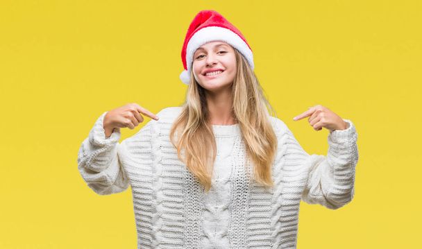 若くてきれいな金髪女性の顔に笑顔と自信を持って、誇りと幸せな指で自分を指している孤立した背景にクリスマスの帽子をかぶって. - 写真・画像