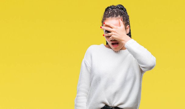 Joven chica afroamericana trenzada de pelo usando suéter de invierno sobre fondo aislado asomándose en shock cubriendo cara y ojos con la mano, mirando a través de los dedos con expresión avergonzada
. - Foto, Imagen