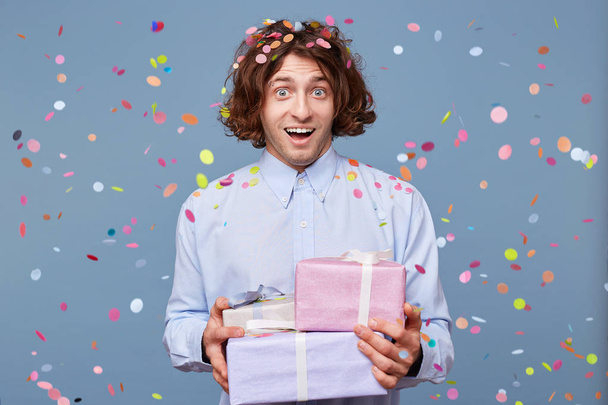 Young man accepts gifts for a new year or birthday, holds three boxes tied up with ribbons, looks happy, confetti fall down isolated on a blue background - Fotografie, Obrázek