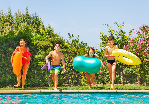 Four happy teens with swim toys standing together on the edge of swimming pool, preparing to jump into the water - Photo, image