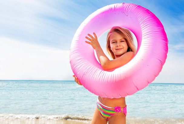 Portrait of beautiful little girl looking through the pink swim ring on the beach - Zdjęcie, obraz