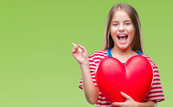 Молода красива дівчина тримає червоне серце над ізольованим тлом дуже щаслива, вказуючи рукою і пальцем на бік
 - Фото, зображення