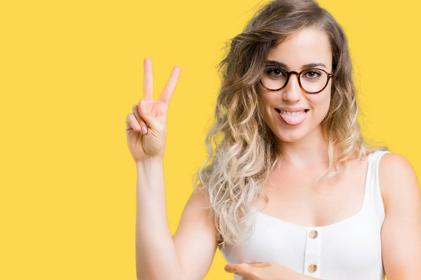 Beautiful young blonde woman wearing glasses over isolated background smiling with happy face winking at the camera doing victory sign. Number two. - Photo, Image