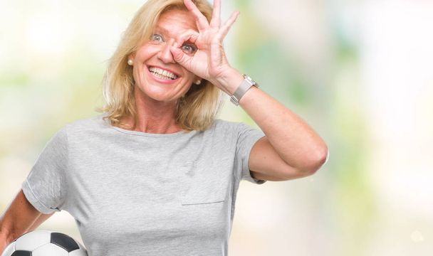 Μέσης ηλικίας ξανθιά γυναίκα που κρατά μπάλα ποδοσφαίρου ποδοσφαίρου πέρα από το απομονωμένο υπόβαθρο με χαρούμενο πρόσωπο χαμογελά κάνει εντάξει σημάδι με το χέρι στο μάτι που αναζητούν μέσα από τα δάχτυλα - Φωτογραφία, εικόνα