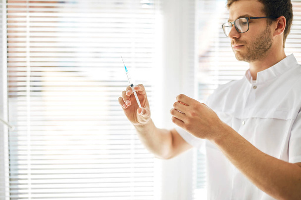 man in medical uniform looking at syringe before injection at workplace - Foto, Imagen
