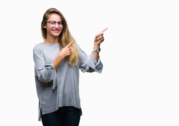 Jovem mulher loira bonita vestindo óculos sobre fundo isolado sorrindo e olhando para a câmera apontando com duas mãos e dedos para o lado
. - Foto, Imagem