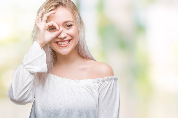 Mladá blond žena izolované pozadí dělá ok gesto rukou, s úsměvem, oko dívá skrze prsty s šťastný obličej. - Fotografie, Obrázek