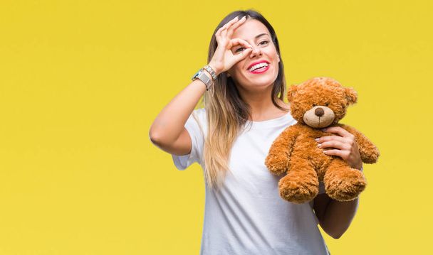 Young beautiful woman holding teddy bear plush over isolated background with happy face smiling doing ok sign with hand on eye looking through fingers - Valokuva, kuva