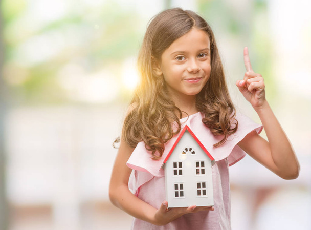 Brunette hispanic girl holding little house surprised with an idea or question pointing finger with happy face, number one - Photo, Image