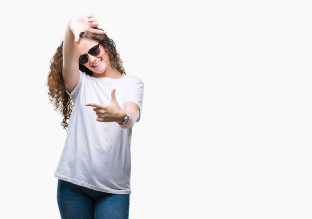Beautiful brunette curly hair young girl wearing sunglasses over isolated background smiling making frame with hands and fingers with happy face. Creativity and photography concept. - Zdjęcie, obraz