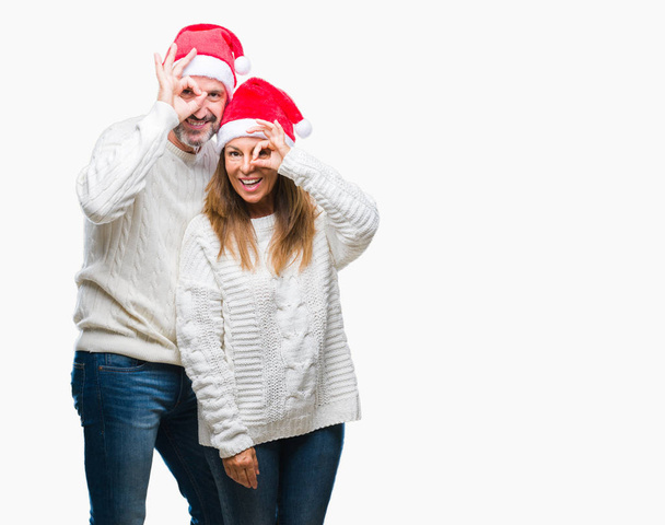 Латиноамериканская пара средних лет в рождественской шляпе на изолированном фоне делает хорошо жест с улыбкой на руке, глаза смотрят сквозь пальцы с счастливым лицом
. - Фото, изображение