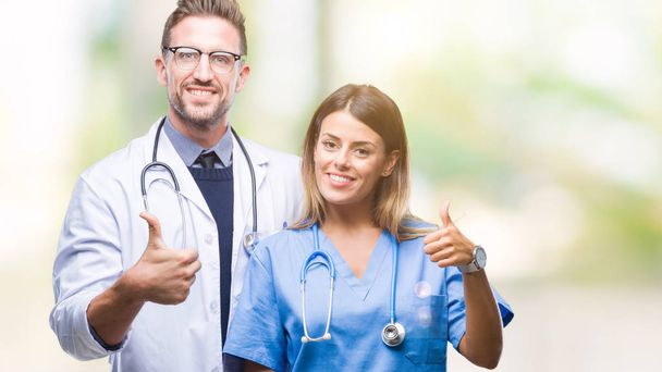 Young couple of doctor and surgeon over isolated background doing happy thumbs up gesture with hand. Approving expression looking at the camera with showing success. - Photo, Image