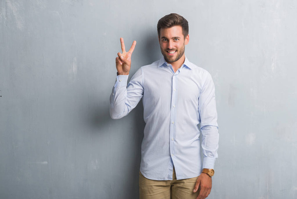 Pohledný mladý obchodník přes zeď šedá grunge elegantní košili ukazující a ukázal prsty číslo dvě, zatímco s úsměvem a spokojené. - Fotografie, Obrázek