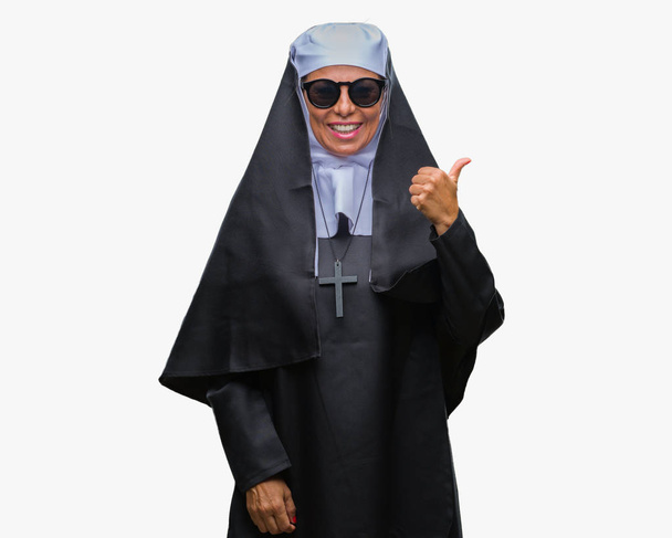 Femme religieuse catholique âgée du Moyen Âge portant des lunettes de soleil sur fond isolé souriant avec le visage heureux regardant et pointant vers le côté avec le pouce vers le haut
. - Photo, image