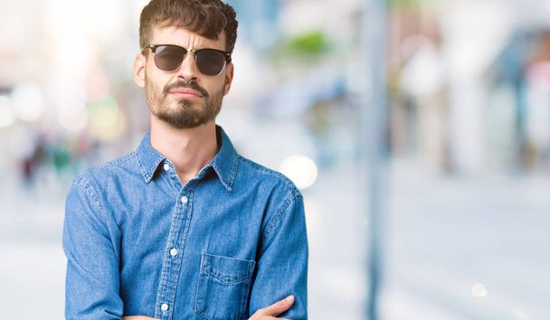 Young handsome man wearing sunglasses over isolated background skeptic and nervous, disapproving expression on face with crossed arms. Negative person. - Photo, Image
