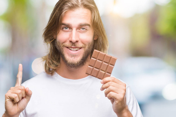 Nuori komea mies, jolla on pitkät hiukset, syö suklaapatukkaa eristetyn taustan päällä yllättynyt ideasta tai kysymyksestä, joka osoittaa sormella onnellisia kasvoja, numero yksi.
 - Valokuva, kuva