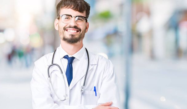 Молодой доктор в больничном пальто на изолированном фоне счастливое лицо со скрещенными руками, смотрящее в камеру. Позитивный человек
. - Фото, изображение