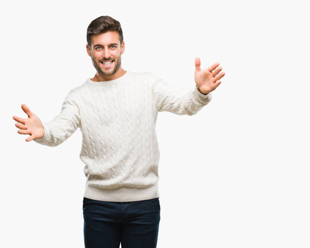 若いハンサムな男はハグするため両手を広げて笑みを浮かべてカメラを見て孤立の背景に冬のセーターを着ています。晴れやかな表情の受け入れ幸せ. - 写真・画像
